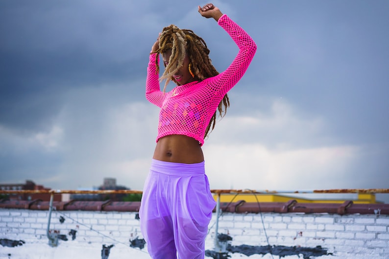 May include: A woman with long dreadlocks dances on a rooftop wearing a pink mesh crop top and purple pants.