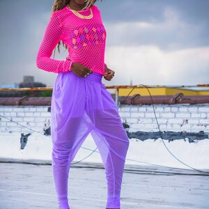 May include: A woman wearing a pink mesh top with a colorful design and purple sheer pants. She is wearing blue platform sandals with a cloud-like pattern.