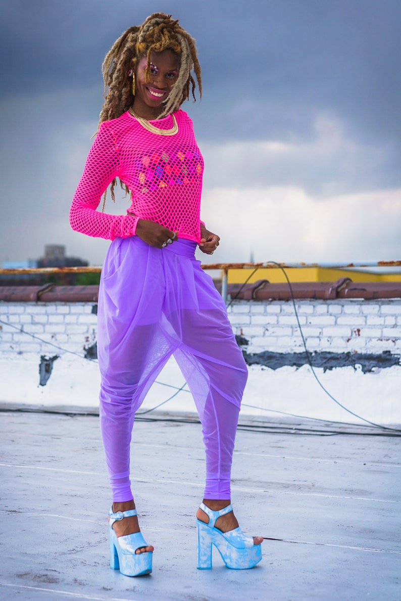 May include: A woman wearing a pink mesh top with a colorful design and purple sheer pants. She is wearing blue platform sandals with a cloud-like pattern.