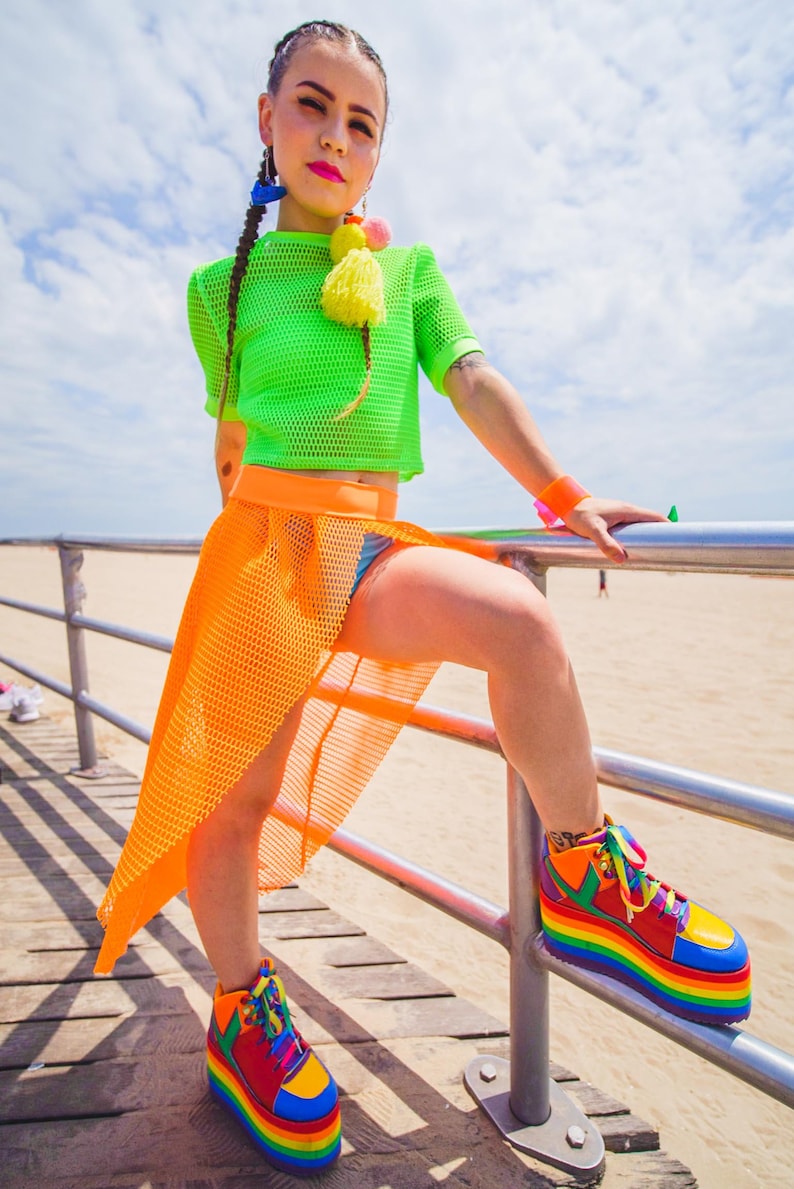 May include: A woman wearing a green mesh crop top, an orange mesh skirt, and rainbow platform sneakers. She is standing on a wooden boardwalk with a metal railing.