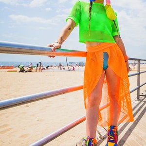 May include: A woman wearing a neon green mesh crop top and an orange mesh skirt. She is standing on a wooden boardwalk with a metal railing in front of her. She is wearing rainbow platform shoes.