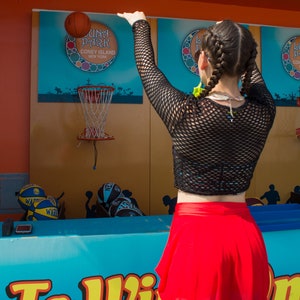 May include: A person wearing a black mesh top and a red skirt throws a basketball at a hoop with the words "Luna Park Coney Island New York" on the backboard. The words "To Win" are visible in the foreground.