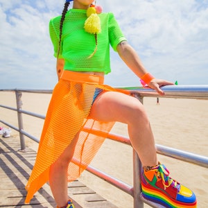 May include: A woman wearing a green mesh crop top, an orange mesh skirt, and rainbow platform sneakers. She is standing on a wooden boardwalk with a metal railing.