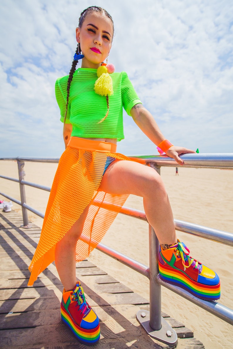 May include: A woman wearing a green mesh crop top, an orange mesh skirt, and rainbow platform sneakers. She is standing on a wooden boardwalk with a metal railing.