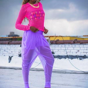 May include: A woman wearing a pink mesh top with a colorful design and purple sheer pants. She is wearing blue platform sandals with a cloud-like pattern.