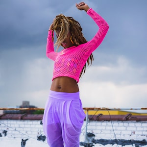 May include: A woman with long dreadlocks dances on a rooftop wearing a pink mesh crop top and purple pants.