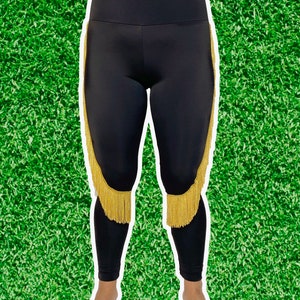 Puede incluir: Leggings negros con ribetes de flecos dorados a los lados. Los leggings son de material elástico y tienen cintura alta.