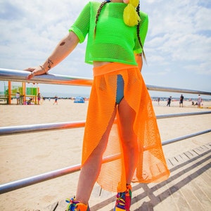 May include: A woman wearing a bright green mesh crop top, an orange mesh skirt, and rainbow platform sneakers. She is standing on a wooden boardwalk with a metal railing in front of her.