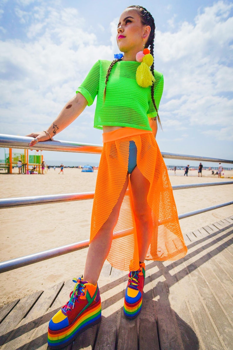 May include: A woman wearing a bright green mesh crop top, an orange mesh skirt, and rainbow platform sneakers. She is standing on a wooden boardwalk with a metal railing in front of her.