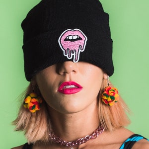 May include: A woman wearing a black beanie with a pink dripping lips patch, a pink and silver chain necklace with a blue strawberry charm, and colourful pom-pom earrings.