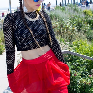 May include: A woman wearing a black fishnet crop top with a gold chain necklace and red sheer trousers. She has long blonde hair in two braids and is wearing blue mirrored sunglasses.