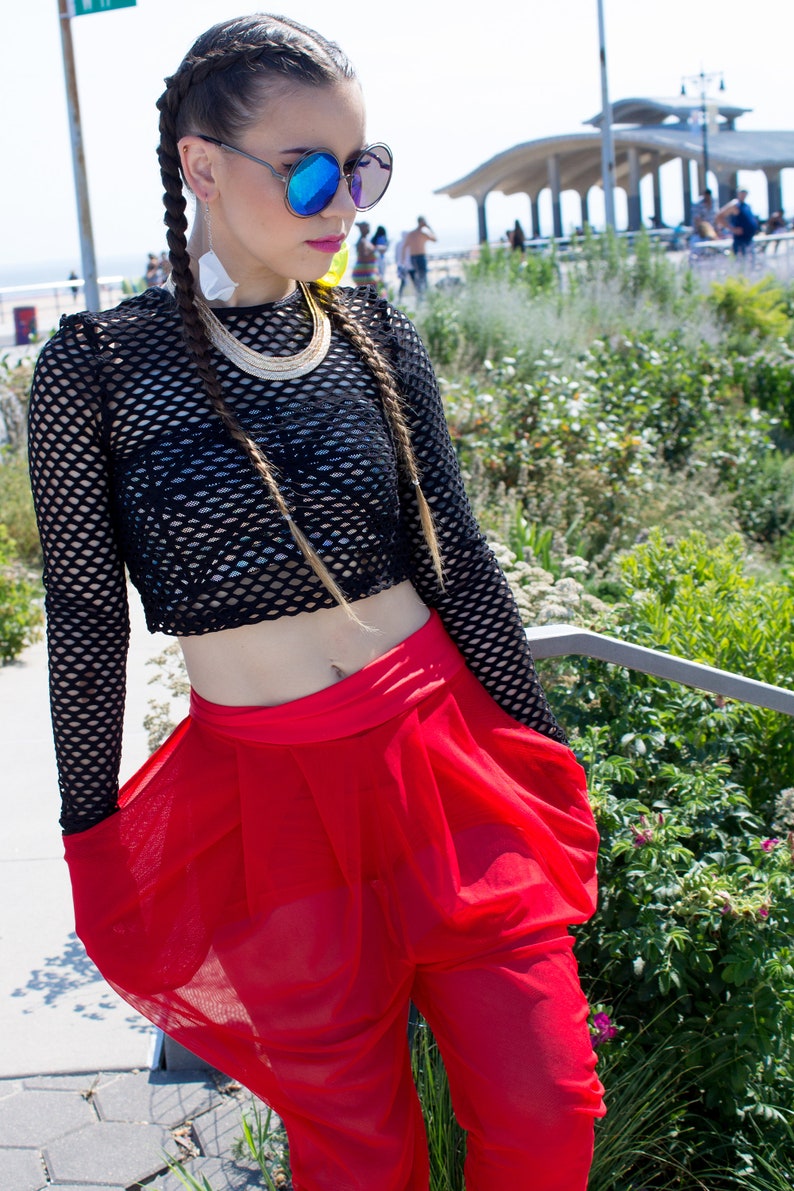 May include: A woman wearing a black fishnet crop top with a gold chain necklace and red sheer trousers. She has long blonde hair in two braids and is wearing blue mirrored sunglasses.