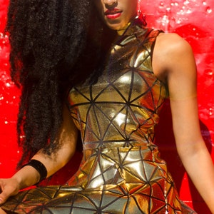 May include: A woman with long black hair wearing a gold metallic sleeveless dress with a geometric pattern. The dress has a high neckline and a fitted bodice. The woman is sitting on a red surface.