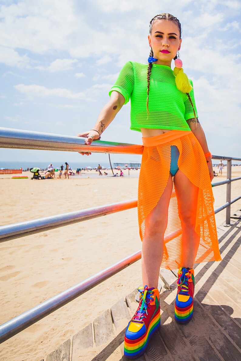May include: A woman wearing a bright green mesh crop top and a bright orange mesh skirt. She is standing on a wooden boardwalk with a beach in the background. She is wearing rainbow platform shoes.