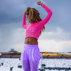 May include: A woman with long dreadlocks dances on a rooftop wearing a pink mesh crop top and purple pants.
