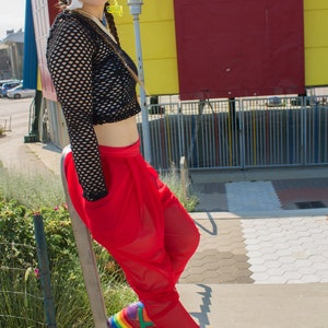 May include: A woman wearing a black fishnet top, red trousers, and rainbow platform trainers. She is standing in front of a colourful wall.