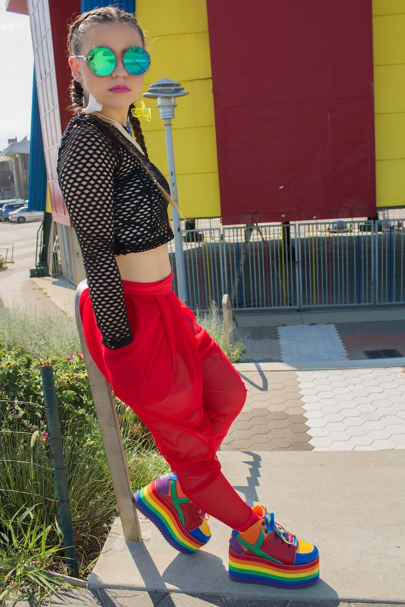 May include: A woman wearing a black fishnet top, red trousers, and rainbow platform trainers. She is standing in front of a colourful wall.