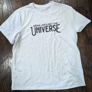 May include: White t-shirt with the text "DRINK AROUND THE UNIVERSE" printed in black. The shirt has short sleeves and a crew neck. The shirt is laying on a dark brown wooden surface.