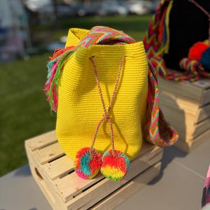 May include: A bright yellow drawstring bag with a colorful woven strap and two matching pom-poms. The bag is sitting on a wooden crate. The pom-poms are a mix of rainbow colors.