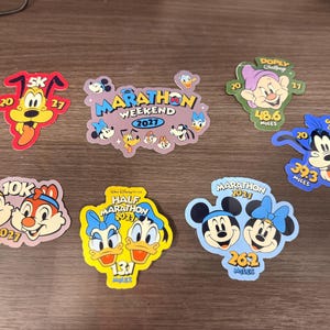 May include: A collection of colorful Disney marathon and race stickers. The stickers feature various characters like Mickey and Minnie Mouse, Goofy, and Chip and Dale, with text indicating race distances and years.