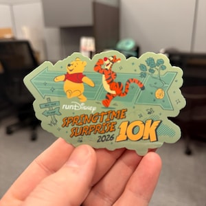 May include: A green sticker featuring Winnie the Pooh and Tigger from Disney's Springtime Surprise 10K run in 2026. The sticker has a light green border with arrow and floral designs. Winnie the Pooh is wearing a red shirt, and Tigger is orange with black stripes.