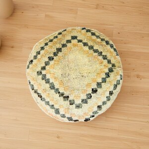 Round Moroccan pouf with a vintage Berber rug pattern in earthy tones, placed on a wooden floor with natural sunlight casting soft shadows.