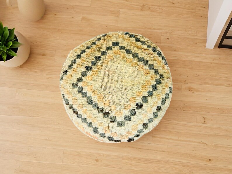Round Moroccan pouf with a vintage Berber rug pattern in earthy tones, placed on a wooden floor with natural sunlight casting soft shadows.