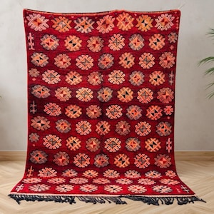 May include: A red rug with a repeating pattern of orange and black geometric shapes. The rug has a fringe along the bottom edge and is displayed against a neutral background. The rug is a decorative textile.
