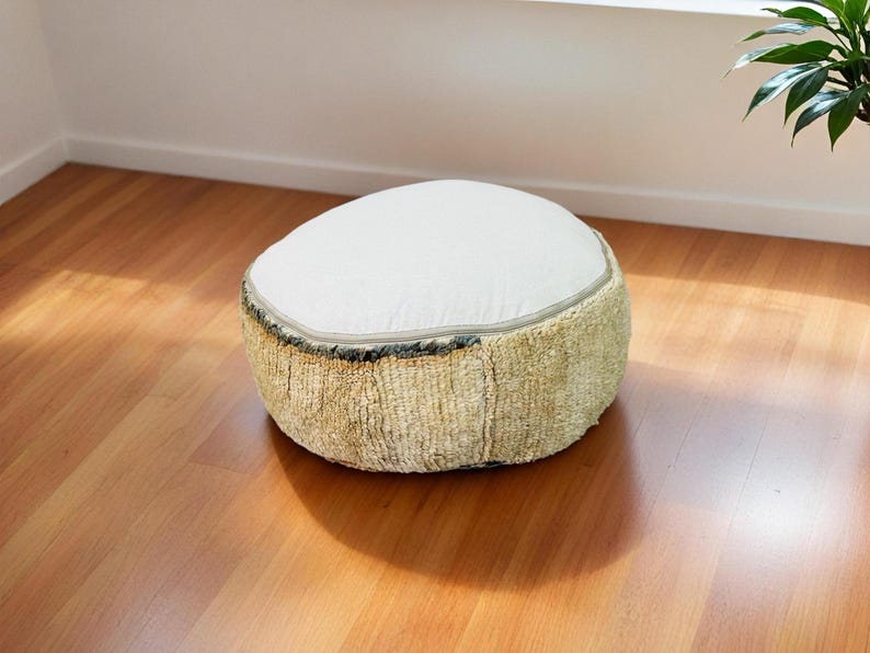 Round Moroccan pouf with a vintage Berber rug pattern in earthy tones, placed on a wooden floor with natural sunlight casting soft shadows.