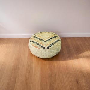 Round Moroccan pouf with a vintage Berber rug pattern in earthy tones, placed on a wooden floor with natural sunlight casting soft shadows.