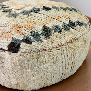 Round Moroccan pouf with a vintage Berber rug pattern in earthy tones, placed on a wooden floor with natural sunlight casting soft shadows.