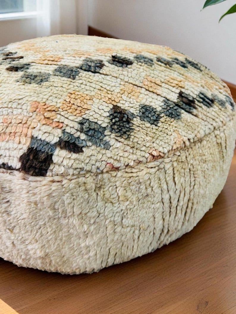 Round Moroccan pouf with a vintage Berber rug pattern in earthy tones, placed on a wooden floor with natural sunlight casting soft shadows.
