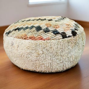 Round Moroccan pouf with a vintage Berber rug pattern in earthy tones, placed on a wooden floor with natural sunlight casting soft shadows.