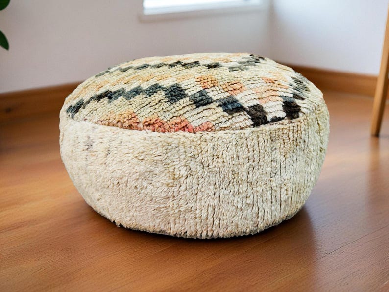 Round Moroccan pouf with a vintage Berber rug pattern in earthy tones, placed on a wooden floor with natural sunlight casting soft shadows.