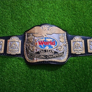 Pode incluir: Um cinto de campeonato de luta de equipas World com uma correia de couro preta e detalhes dourados e prateados. A placa central apresenta uma águia e a palavra "World". O cinto está sobre uma superfície verde.