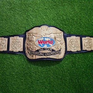 Può includere: Una cintura da campionato di wrestling in pelle nera con una grande placca centrale dorata raffigurante un'aquila, un globo blu e la scritta World Tag Team Wrestling Champions, affiancata da quattro piccole placche laterali quadrate dorate.