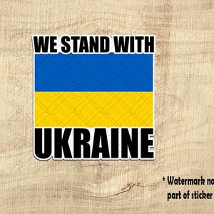 May include: A sticker with the text "WE STAND WITH UKRAINE" in bold black letters. The sticker features the Ukrainian flag, with a blue stripe over a yellow stripe. The background is a light brown wood grain.