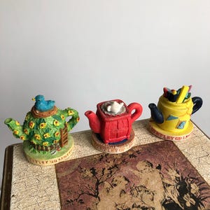 May include: Three decorative teapots with unique designs. One is green with a bird on top, another is red with a white cup, and the third is yellow with pencils. Each teapot sits on a small, round base with text. The teapots are on a decorative surface.