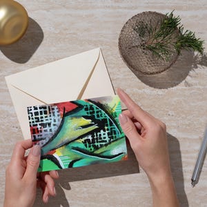 Könnte beinhalten: Eine Grußkarte mit einem abstrakten Design in Grün, Rot und Gelb wird von Händen gehalten. Ein cremefarbener Umschlag, ein goldener Schmuck, eine kleine Glasschale mit Grün und ein silberner Stift sind ebenfalls sichtbar.