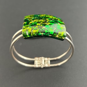 Könnte beinhalten: Ein Armreif aus silberfarbenem Metall mit einem gewölbten, rechteckigen Mittelstück aus geschmolzenem Glas. Das Glaselement zeigt ein lebhaftes, gesprenkeltes Muster in Smaragdgrün, Limettengrün und Goldtönen mit einer glänzenden Oberfläche.