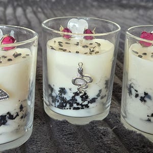 Peut inclure: Trois bougies en verre avec de la cire crème, décorées de fleurs séchées, de breloques et de mouchetures noires. Une bougie a un charme de papillon de nuit, une autre un serpent et la dernière un soleil. Les bougies sont dans des supports en verre transparent.