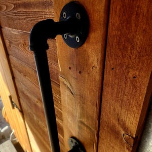 May include: Black pipe handle attached to a wooden door. The handle is made of black metal pipes and fittings, mounted with black circular flanges. The door is made of stained wooden planks.
