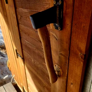 May include: A black axe head mounted on a wooden door. The axe is attached to a wooden handle. The door is made of stained wood and has a metal hinge and a small metal plate.
