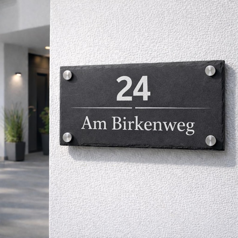 Op de afbeelding: Een rechthoekig huisnummerbord van zwarte leisteen met het nummer 24 en de tekst Am Birkenweg in witte letters. Het bord is bevestigd met vier ronde, zilverkleurige metalen afstandhouders op de hoeken voor een strakke uitstraling.