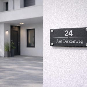 Op de afbeelding: Een rechthoekig huisnummerbord van zwarte leisteen met het nummer 24 en de tekst Am Birkenweg in witte letters. Het bord is bevestigd met vier ronde, zilverkleurige metalen afstandhouders op een gestructureerde witte muur.