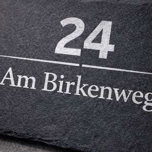Op de afbeelding: Een rechthoekig huisnummerbord van donkergrijze leisteen met het getal 24 in een vet wit schreefloos lettertype boven een dunne horizontale witte lijn, met de tekst Am Birkenweg in het wit onder de lijn in hetzelfde lettertype.
