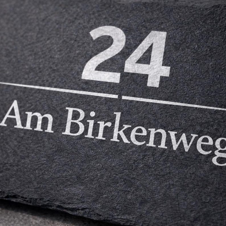 Op de afbeelding: Een rechthoekig huisnummerbord van donkergrijze leisteen met het getal 24 in een vet wit schreefloos lettertype boven een dunne horizontale witte lijn, met de tekst Am Birkenweg in het wit onder de lijn in hetzelfde lettertype.