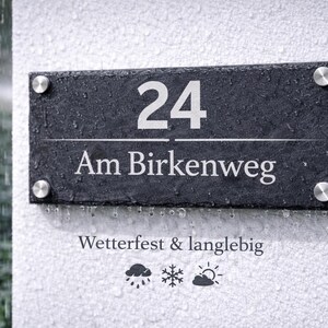 Op de afbeelding: Een rechthoekig huisnummerbord van zwarte leisteen met het nummer 24 en de tekst Am Birkenweg in wit gegraveerd. Het bord is bevestigd met vier zilverkleurige metalen afstandhouders en bevat de tekst Wetterfest & langlebig met weericonen.