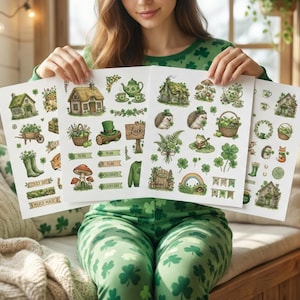 May include: A person holding three sheets of stickers with St. Patrick's Day themed illustrations. The stickers feature houses, clovers, mushrooms, and other green and brown elements. The person is wearing green pajamas with shamrock patterns.