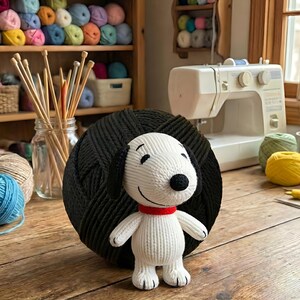Może przedstawiać: Ręcznie robiona biało-czarna pluszowa zabawka Snoopy z czerwoną obrożą stoi przed dużą czarną kulą przędzy. Druty do robienia na drutach i przędza są widoczne w tle, na drewnianej powierzchni.